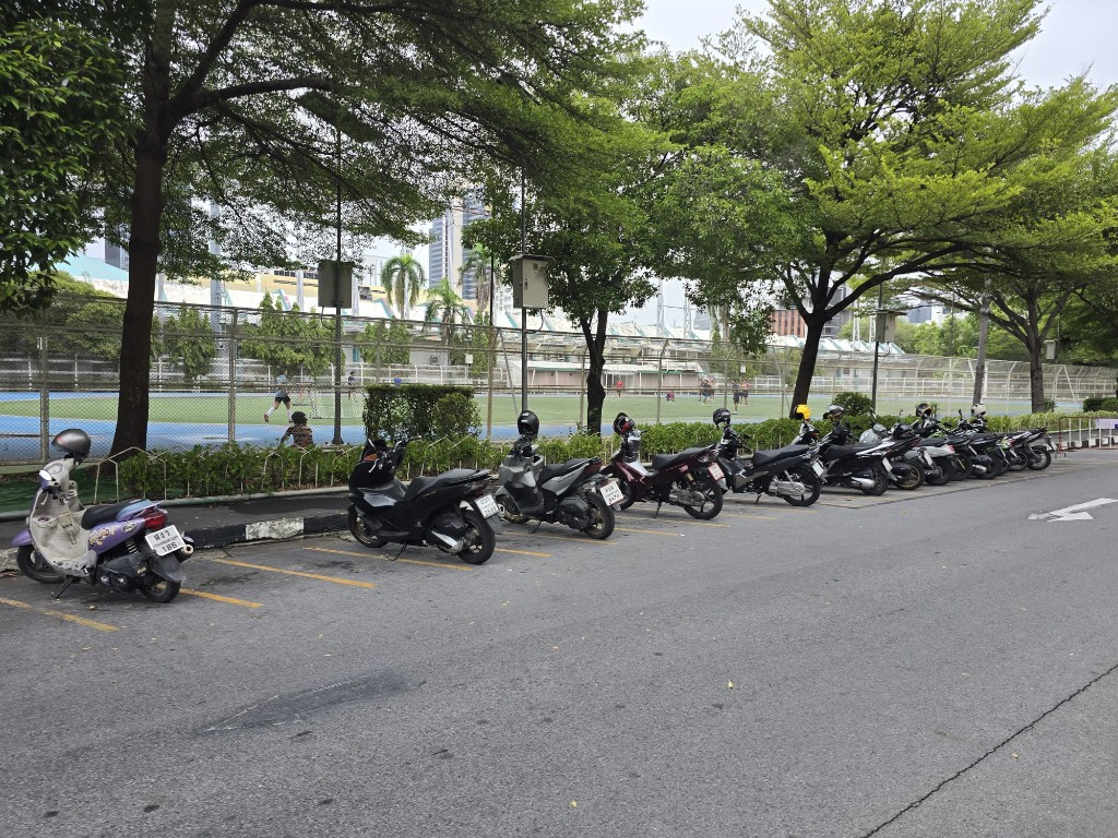 Parked scooters along a tree-lined street beside a blue sports court